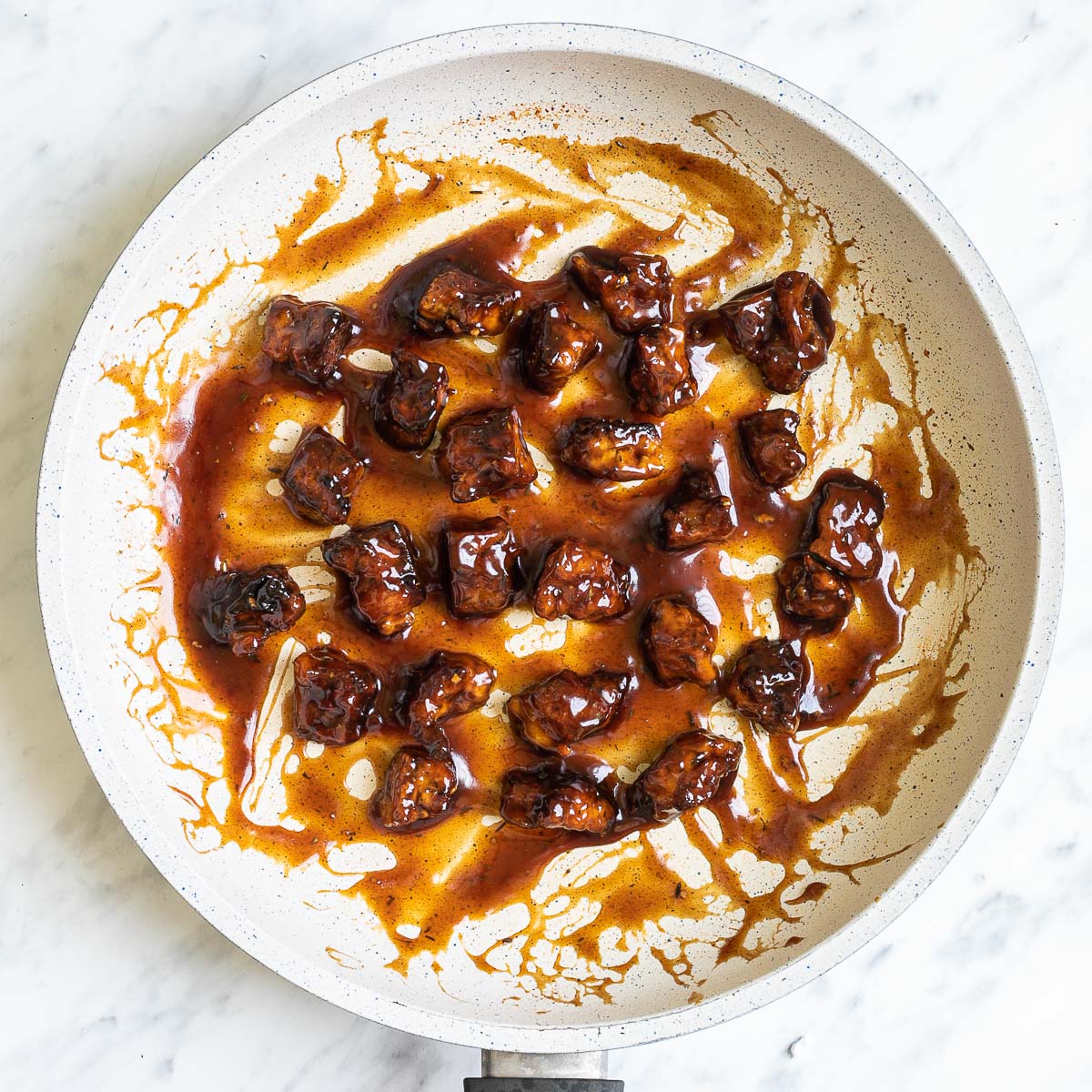 Brown sticky tofu pieces in a white frying pan.