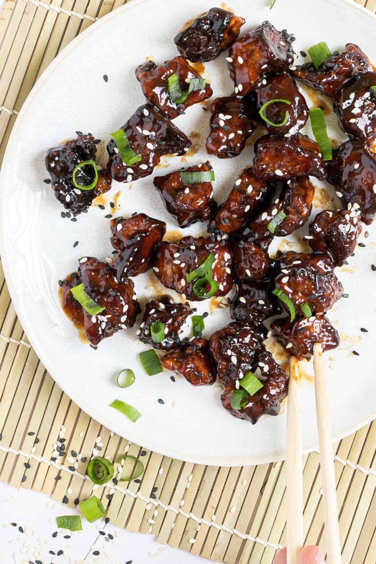 Dark brown sticky juice tofu pieces on a white plate sprinkled with black and white sesame seeds and chopped scallion. The end of a chopsticks is about to grab a piece.