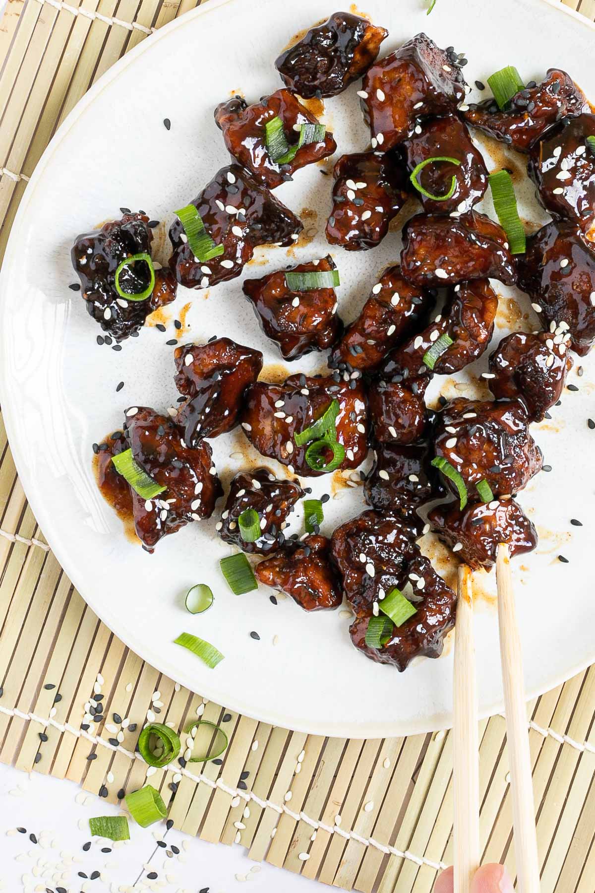 Dark brown sticky juice tofu pieces on a white plate sprinkled with black and white sesame seeds and chopped scallion. The end of a chopsticks is about to grab a piece. 