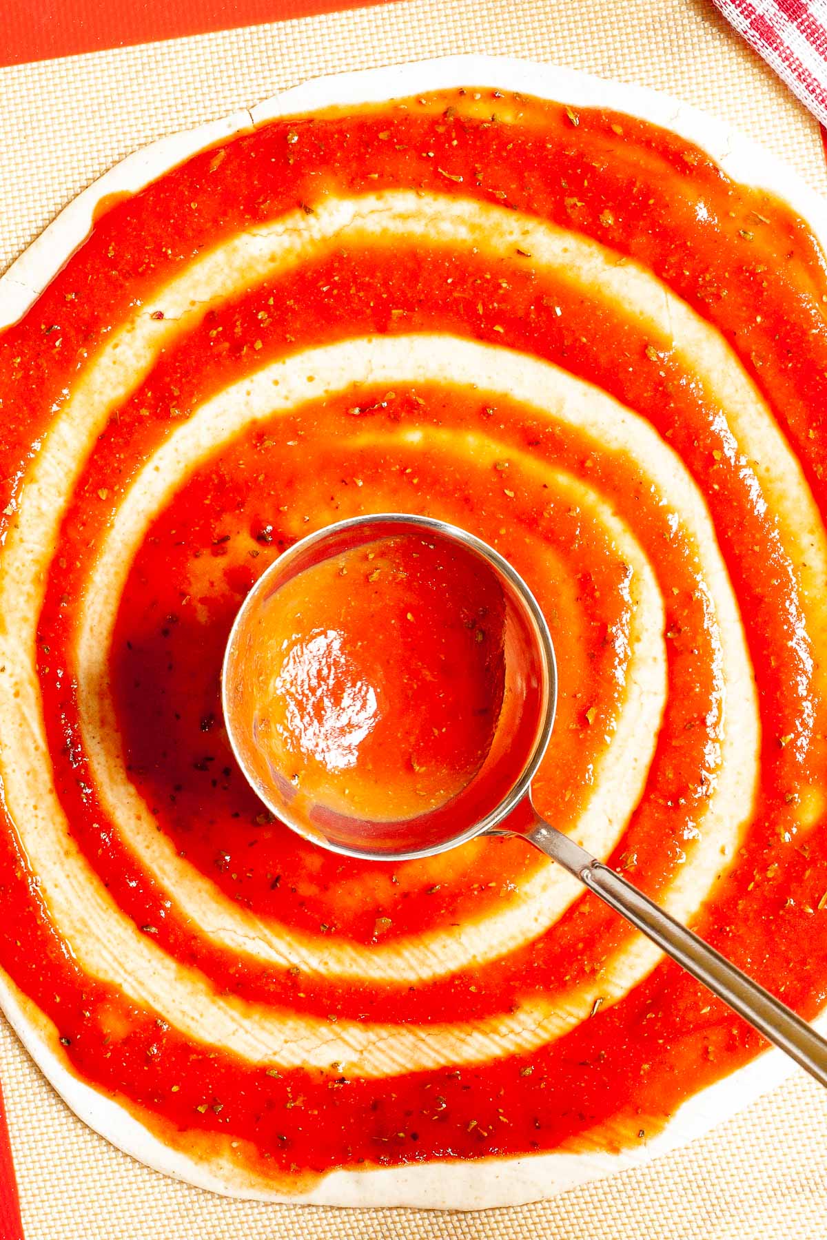 Red pizza sauce is spread in circles on top of a pizza crust with a ladle.