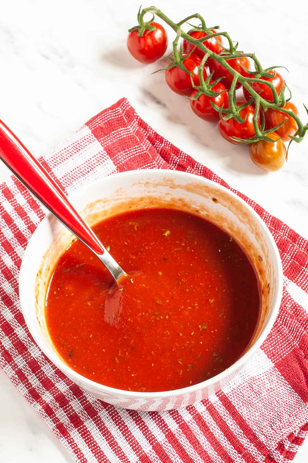Red pizza sauce in a white bowl with a spoon. Red cherry tomato vine is in the background