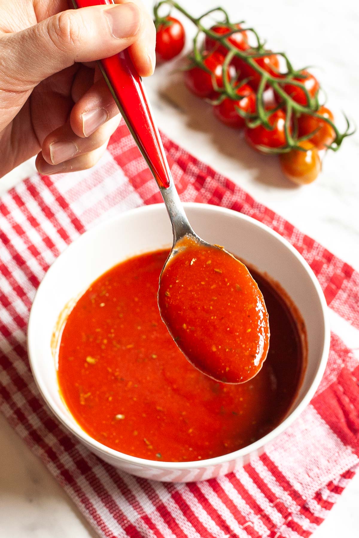 Red pizza sauce in a white bowl. A hand is holding a spoon taking some of the sauce closer. Red cherry tomato vine is in the background