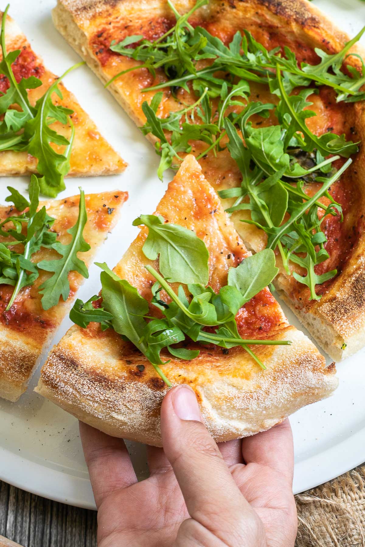 A pizza cut into slices with fluffy crispy crust topped with tomato sauce, melted cheese and fresh arugula on a white plate. A hand is taking one slice.