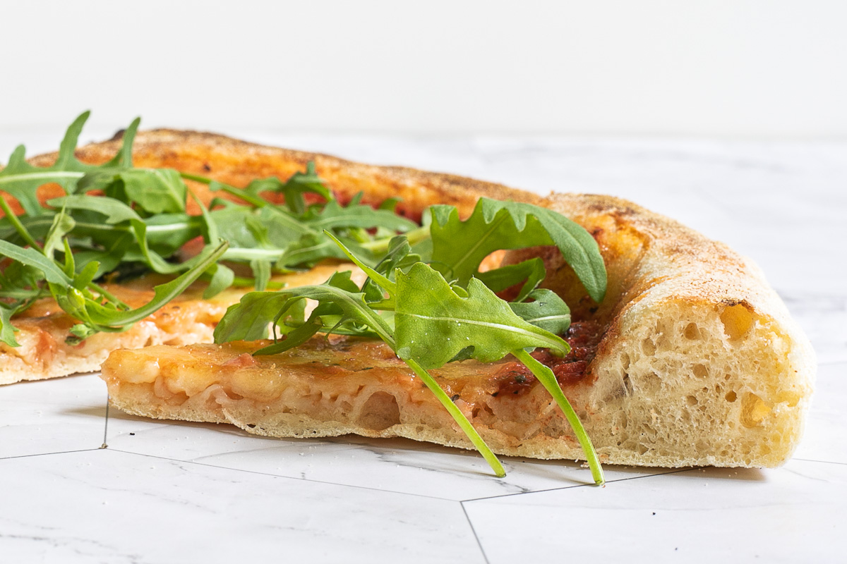A slice of tomato based pizza topped with fresh arugula from the side to see the airy rim.