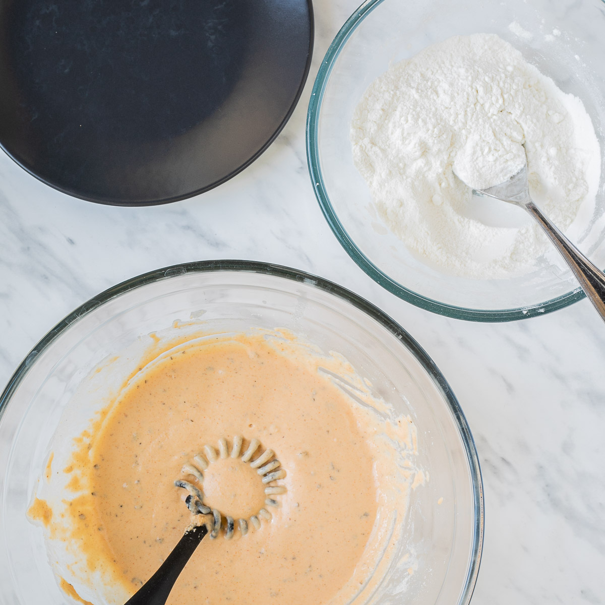 An empty black plate, a glass bowl with white flour, and a glass bowl with orange wet batter and a whisk.