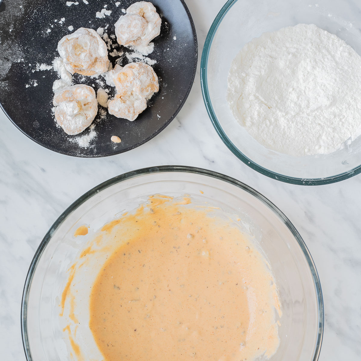 A black plate with breaded cauliflower, a glass bowl with white flour, and a glass bowl with orange wet batter.