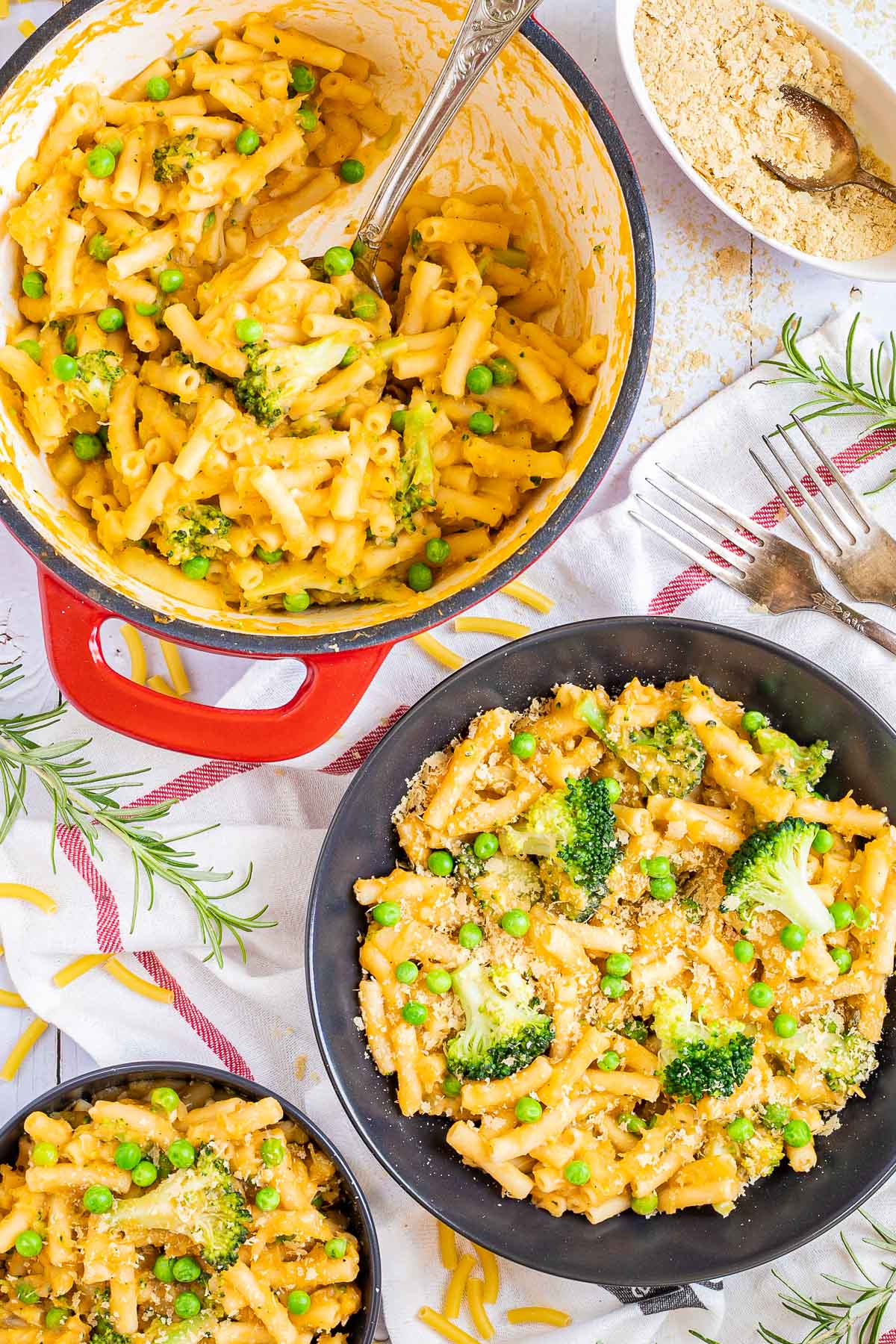 A red white enameled Dutch oven and 2 black bowls are serving short pasta in thick yellow sauce with broccoli florets and green peas added as toppings.