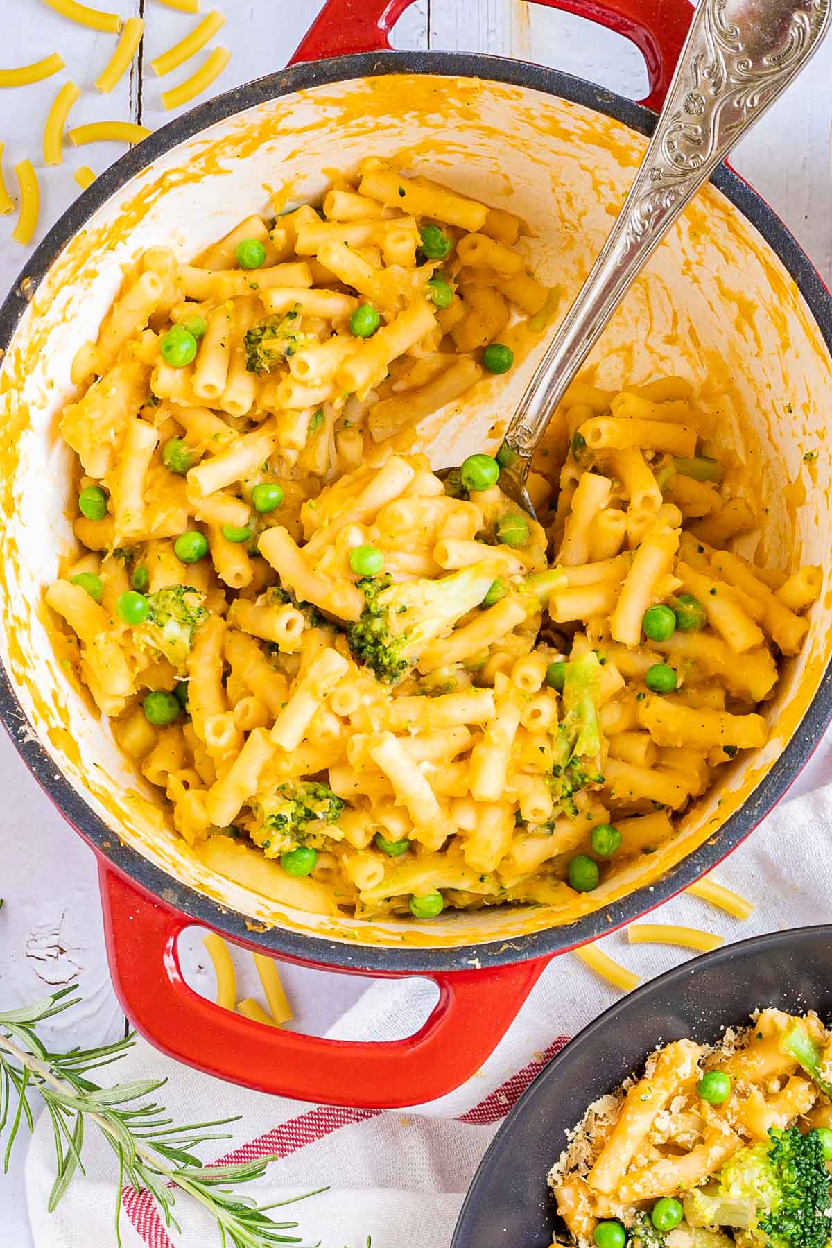 A red white enameled Dutch oven and 1 black bowl are serving short pasta in thick yellow sauce with broccoli florets and green peas added as toppings.