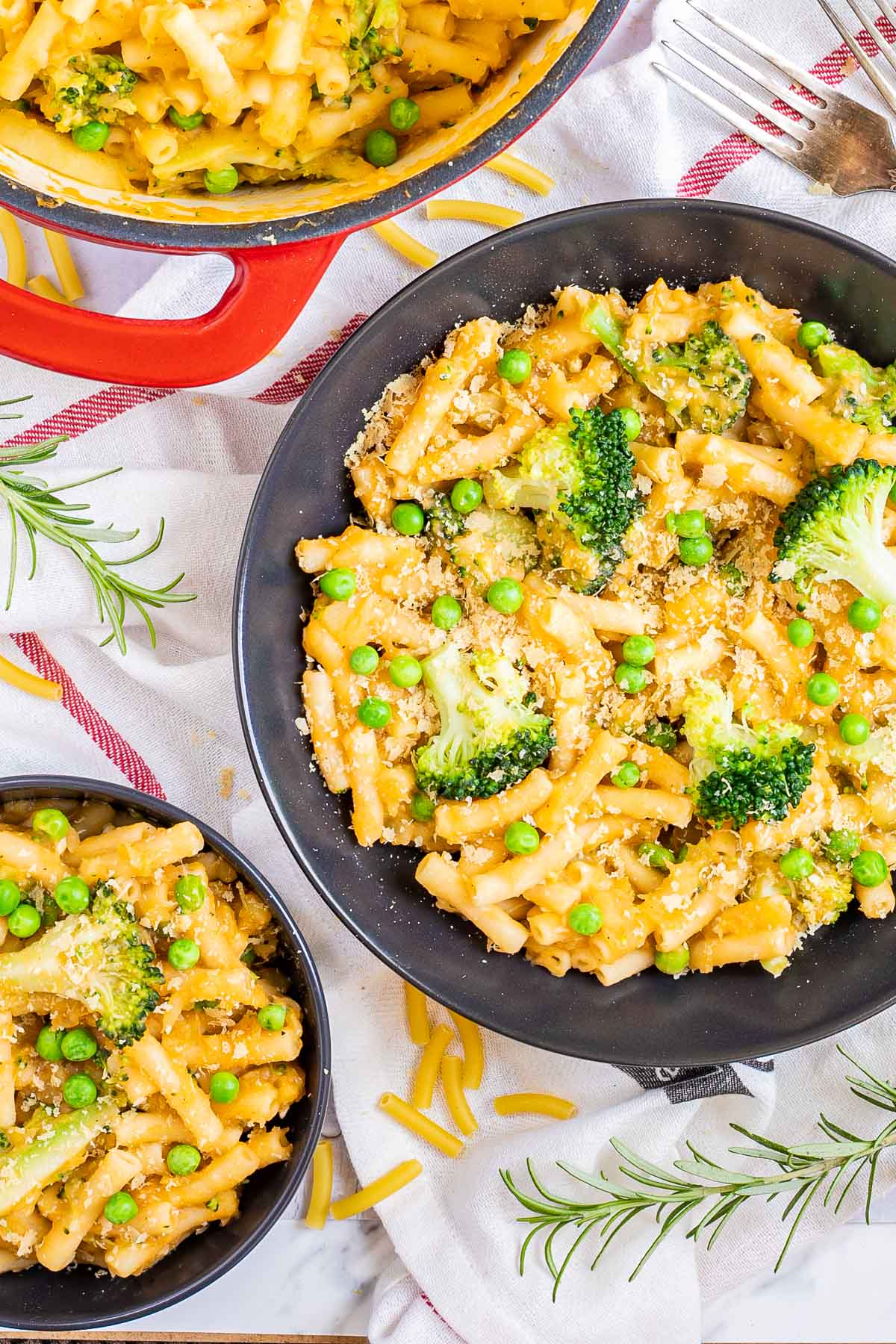 A red white enameled Dutch oven and 2 black bowls are serving short pasta in thick yellow sauce with broccoli florets and green peas added as toppings.