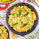 A red white enameled Dutch oven and 2 black bowls are serving short pasta in thick yellow sauce with broccoli florets and green peas added as toppings.
