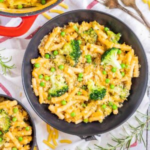 A red white enameled Dutch oven and 2 black bowls are serving short pasta in thick yellow sauce with broccoli florets and green peas added as toppings.