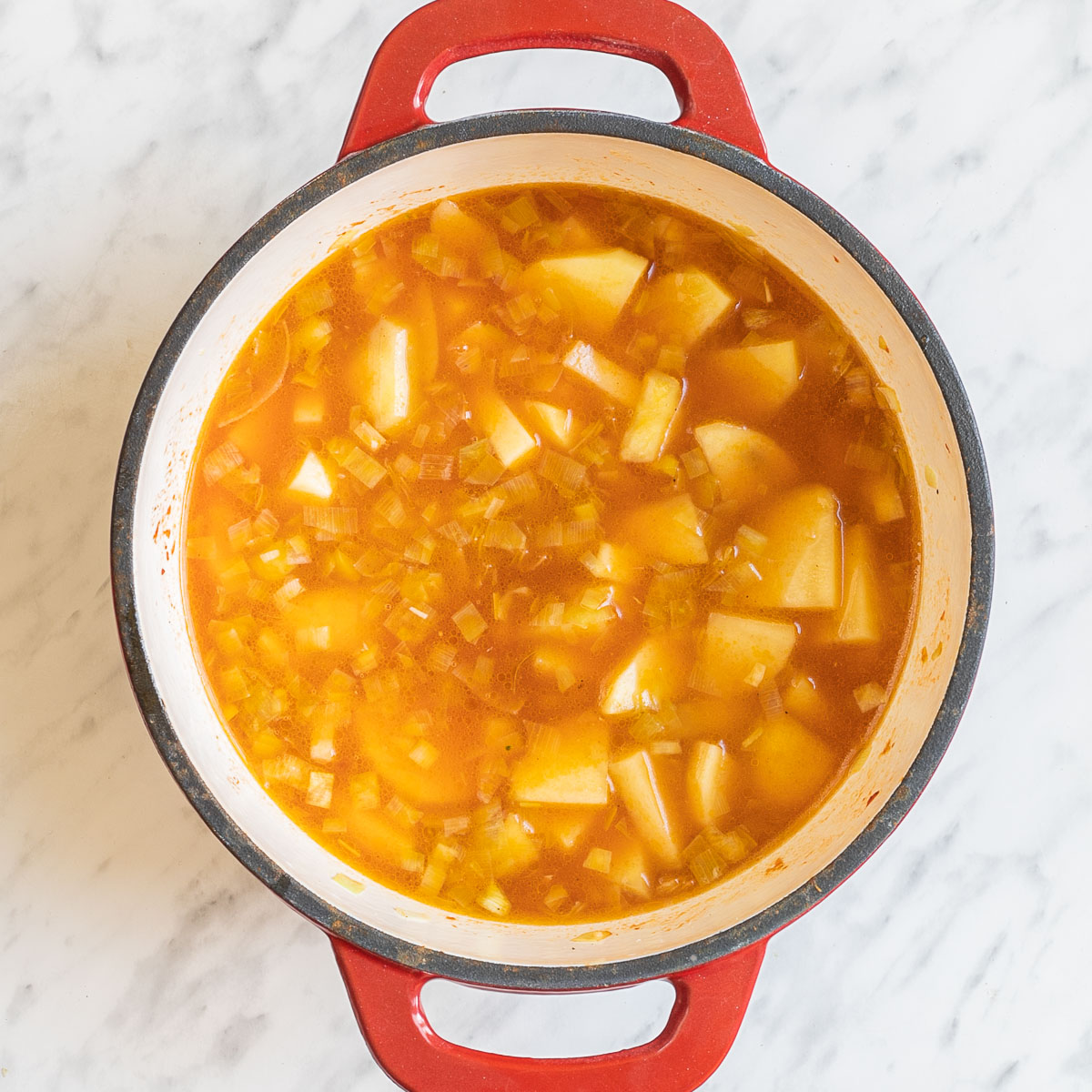 A red white enameled Dutch oven with large potato cubes and onion pieces in an orange liquid.
