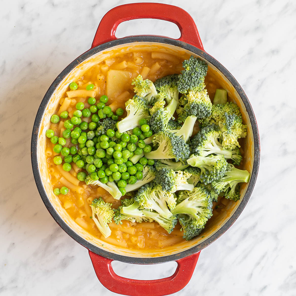 A red white enameled Dutch oven with thick stew-like orange sauce topped with broccoli florets and green peas.