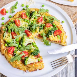 Hash brown crust pizza with topped with tomato sauce, broccoli, peas cherry tomatoes, arugula, and shredded melted cheese. Served on a white plate. A slice is missing and a fork and knife are placed instead there.
