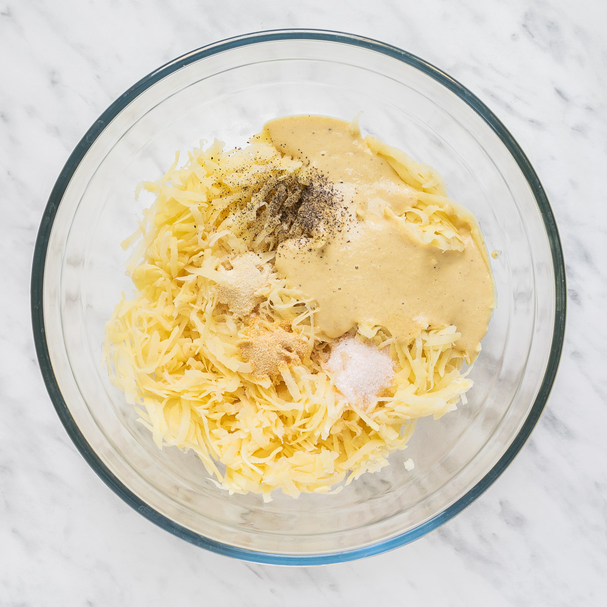Shredded potato, spices, and light brown liquid in a glass bowl.