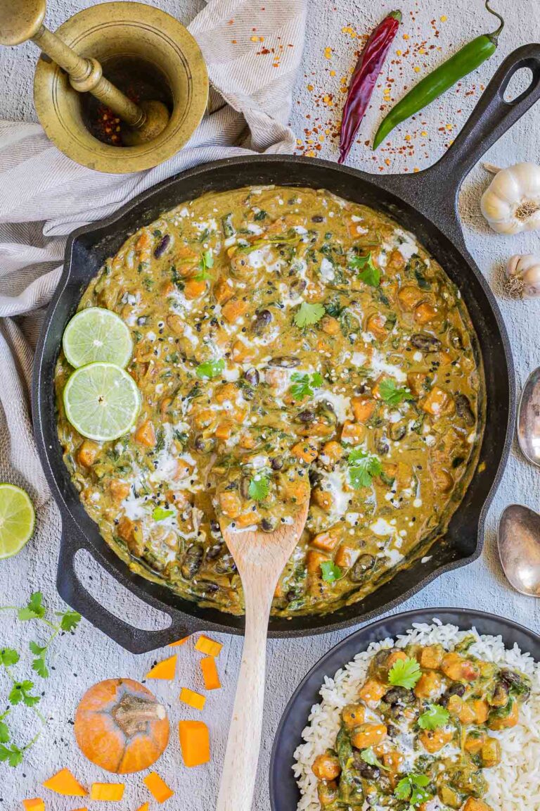 Cast iron skillet with a green-brown curry sauce, orange butternut squash cubes, red kidney beans, and spinach pieces. It is drizzled with white sauce and topped with fresh green herbs. A small bowl with rice and curry is next to it.