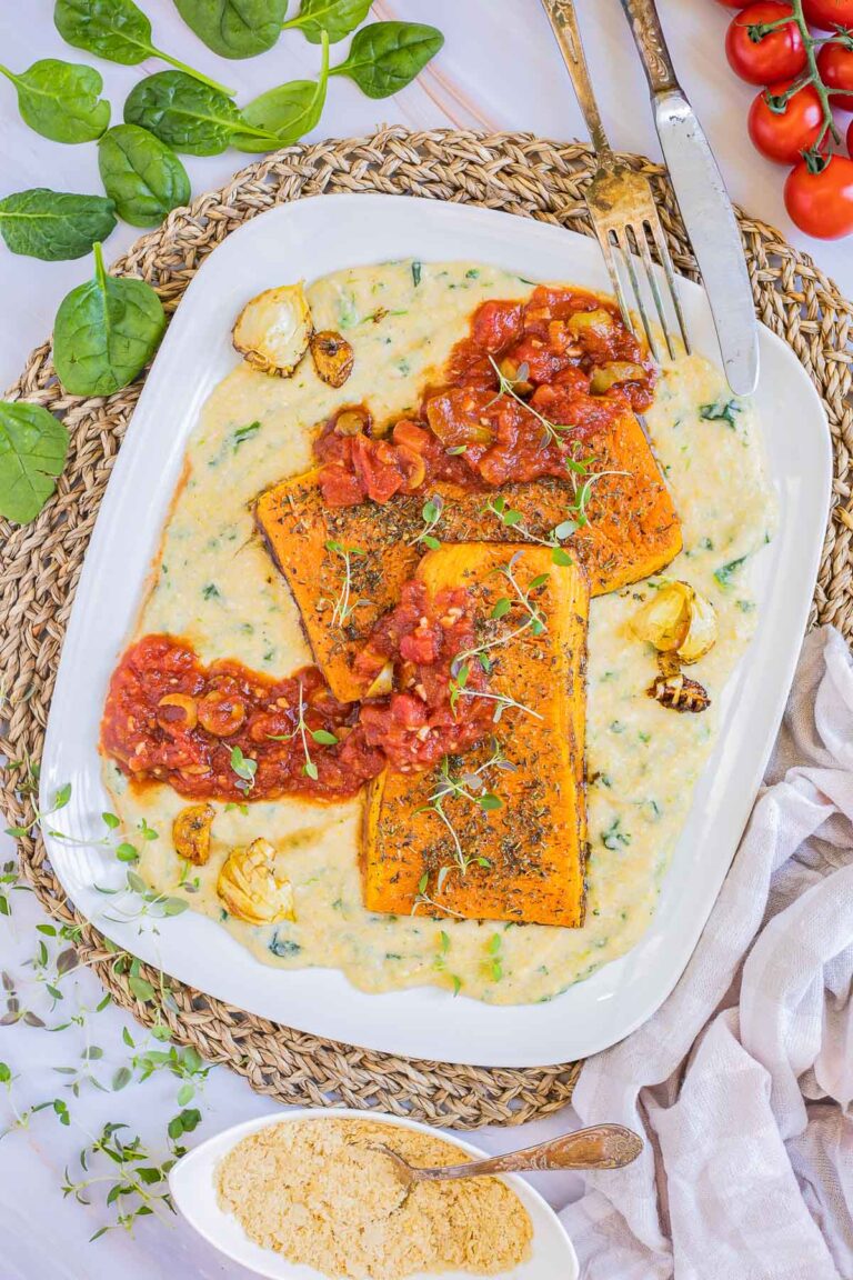 Large white plate with creamy polenta and large butternut squash slices drizzled with red tomato sauce and topped with fresh herbs.