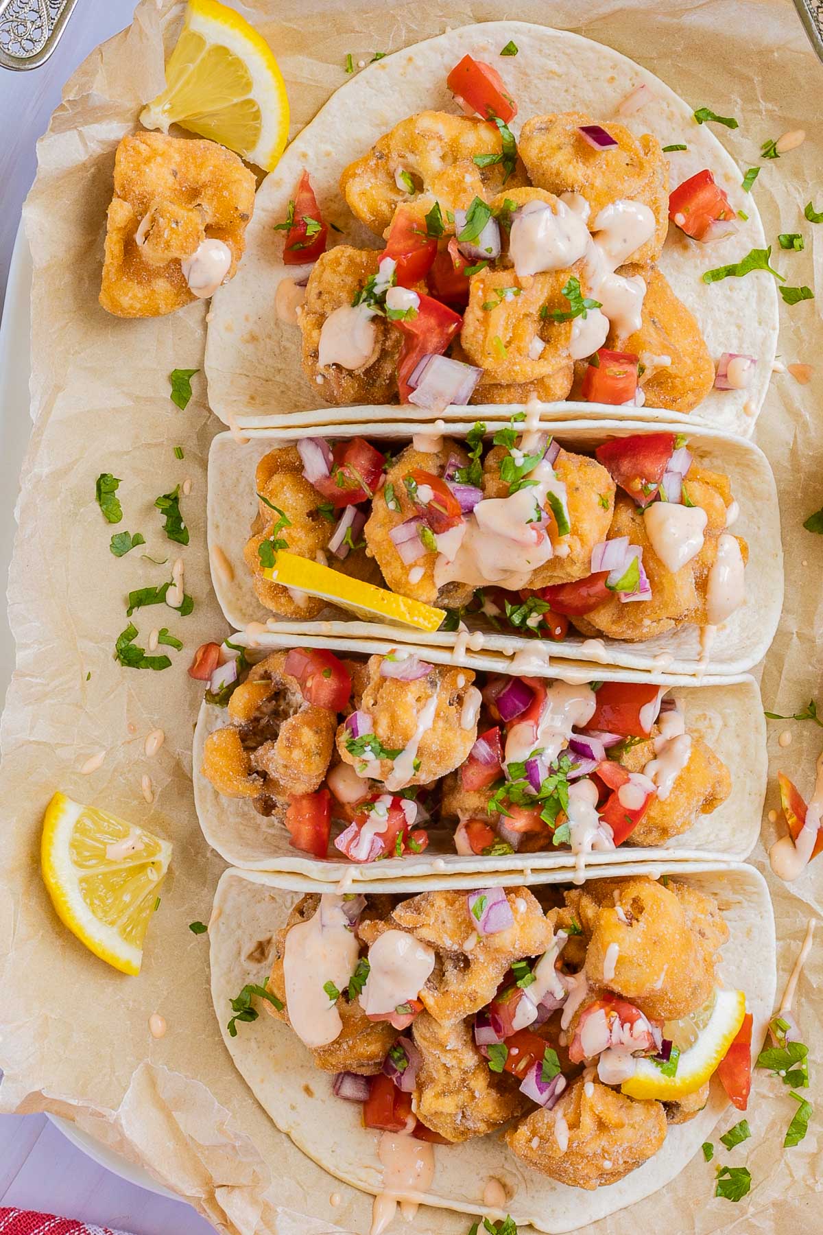 4 small tortillas folded in half on parchment paper filled with cauliflower florets, tomato, and onion pieces drizzled with a light pink sauce. Lemon wedges are next to them.