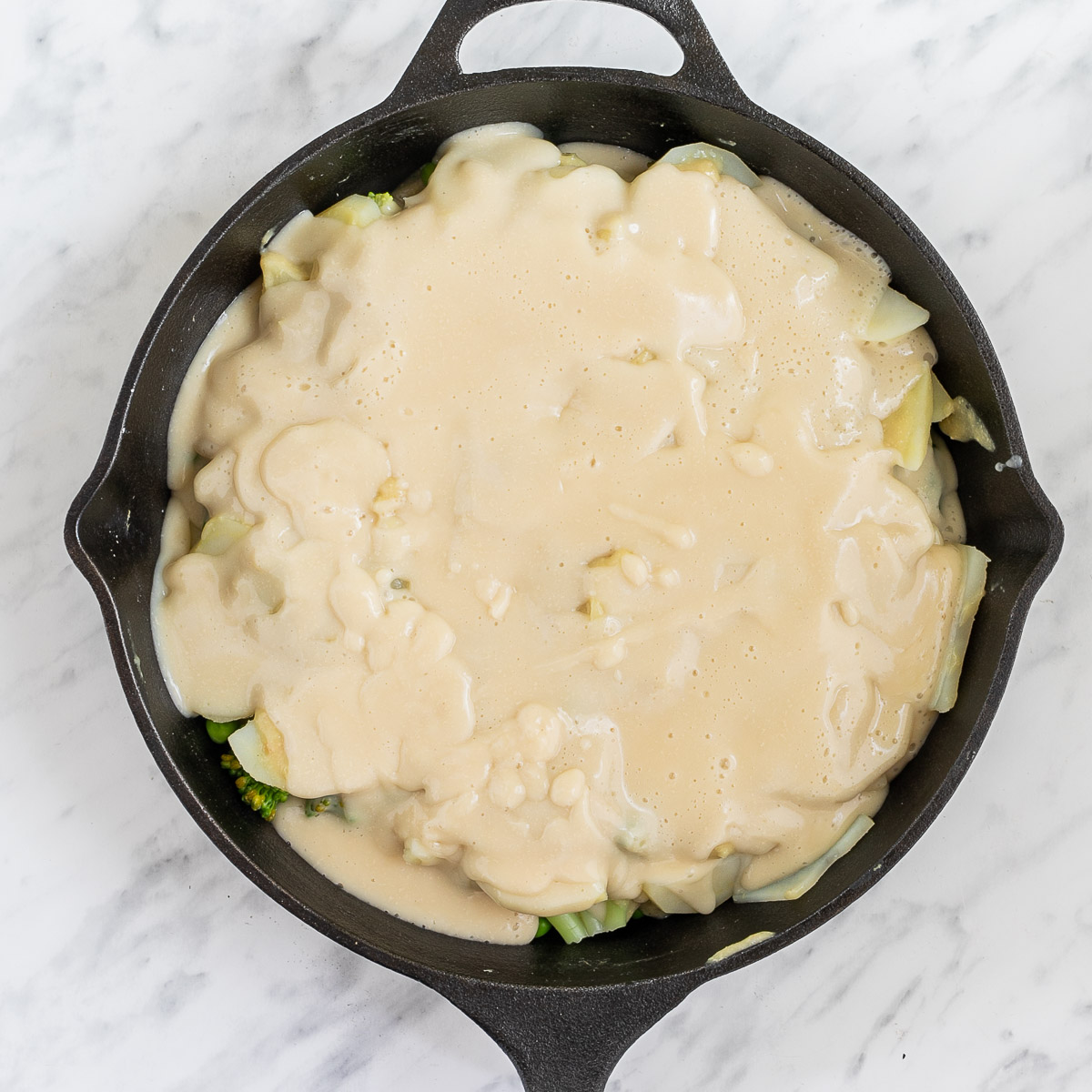 Black cast iron skillet with 2-3 layers of potato slices, broccoli, and green peas. topped with a yellow thick sauce.