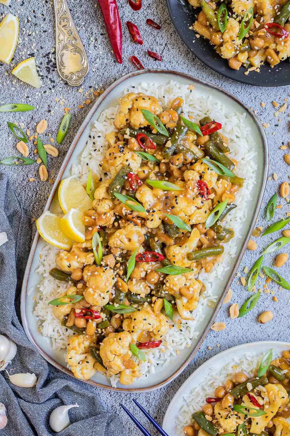 Large oval blue plate with white rice light brown sticky cauliflower, green beans, red chili peppers, scallion and black sesame seeds. Other portions are in side dishes and many ingredients are scattered around. 