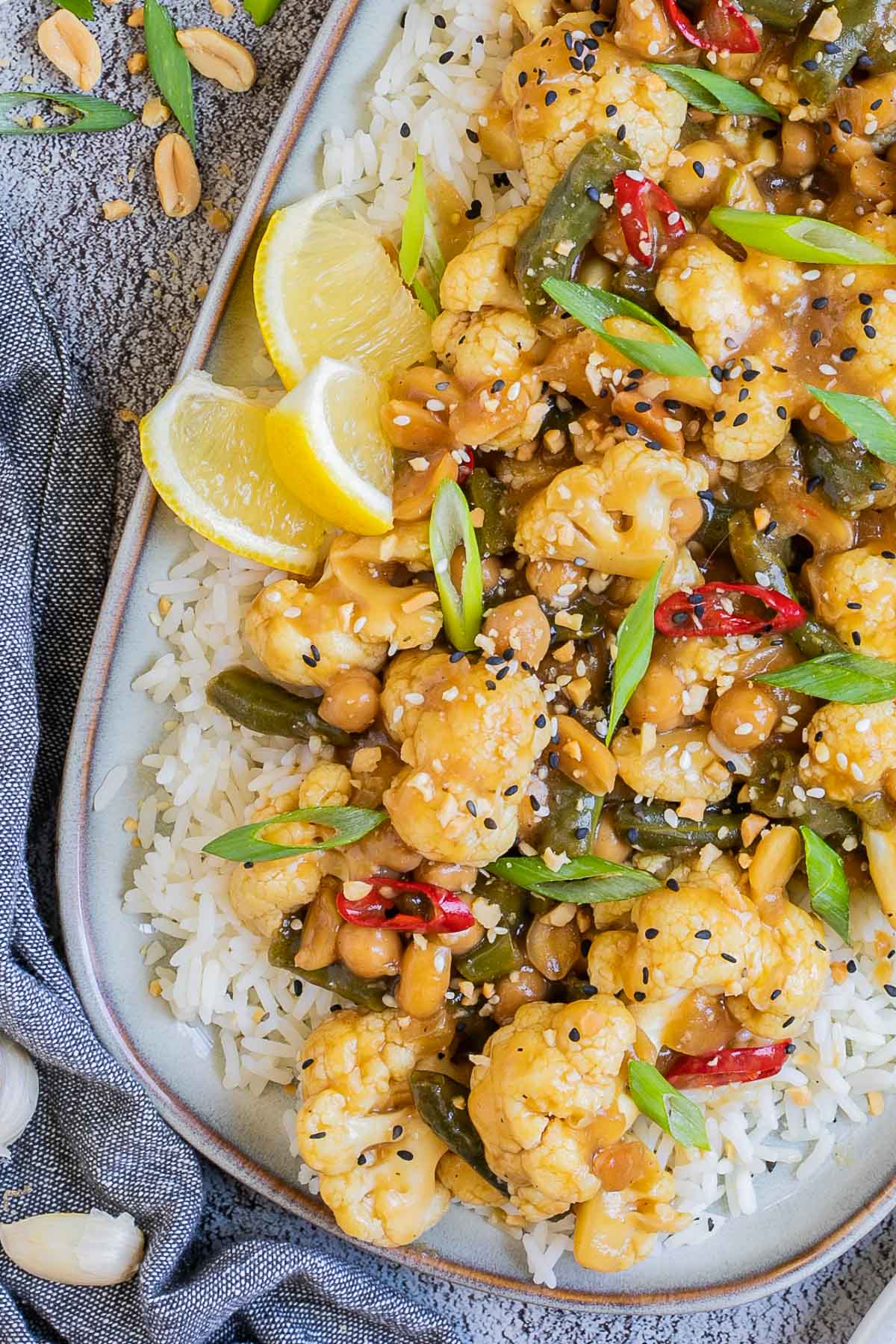 Large oval blue plate with white rice light brown sticky cauliflower, green beans, red chili peppers, scallion and black sesame seeds. Other portions are in side dishes and many ingredients are scattered around.