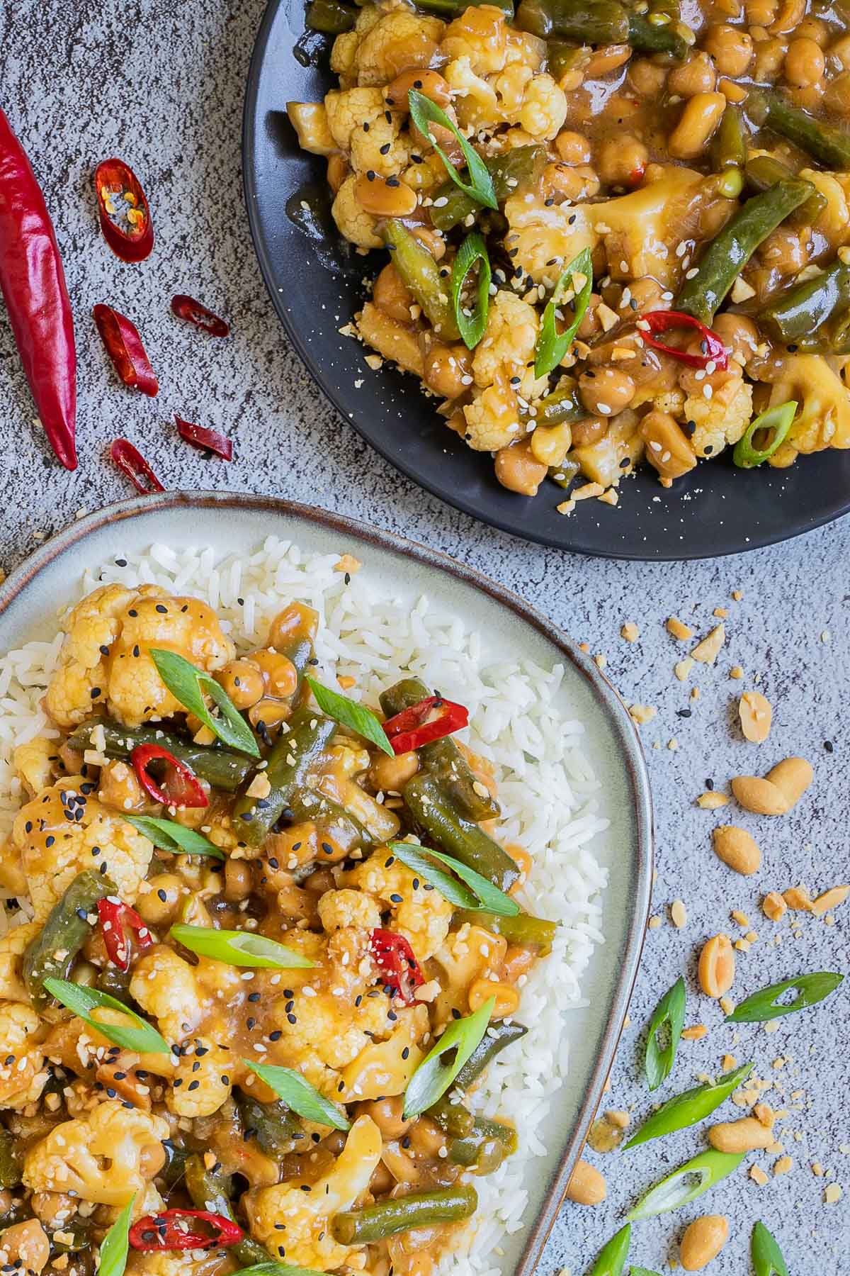 Large oval blue plate with white rice light brown sticky cauliflower, green beans, red chili peppers, scallion and black sesame seeds. Other portions are in side dishes and many ingredients are scattered around.