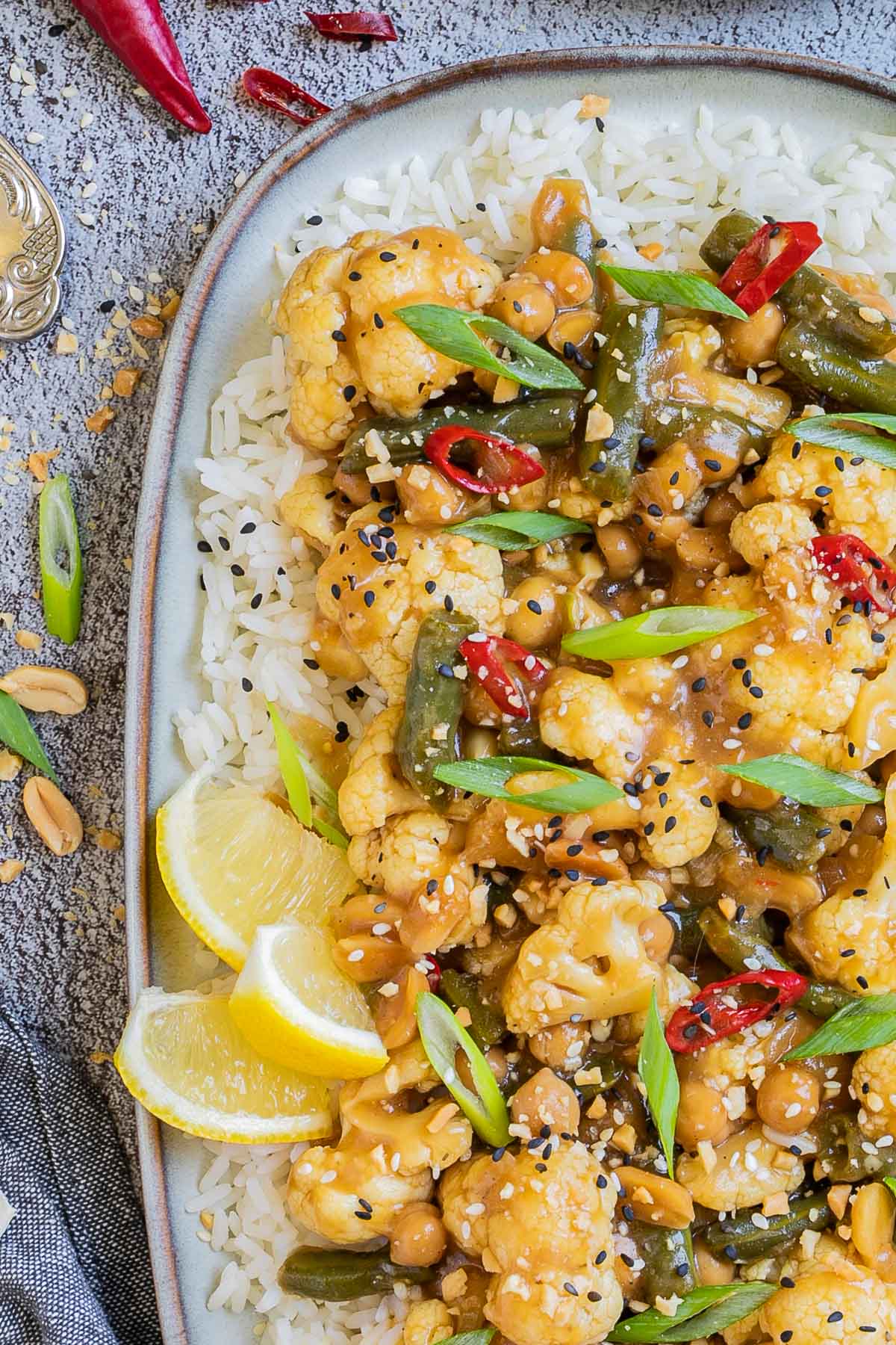Large oval blue plate with white rice light brown sticky cauliflower, green beans, red chili peppers, scallion and black sesame seeds. Other portions are in side dishes and many ingredients are scattered around.