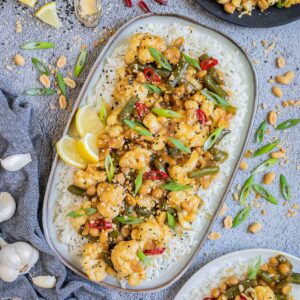 Large oval blue plate with white rice light brown sticky cauliflower, green beans, red chili peppers, scallion and black sesame seeds. Other portions are in side dishes and many ingredients are scattered around.