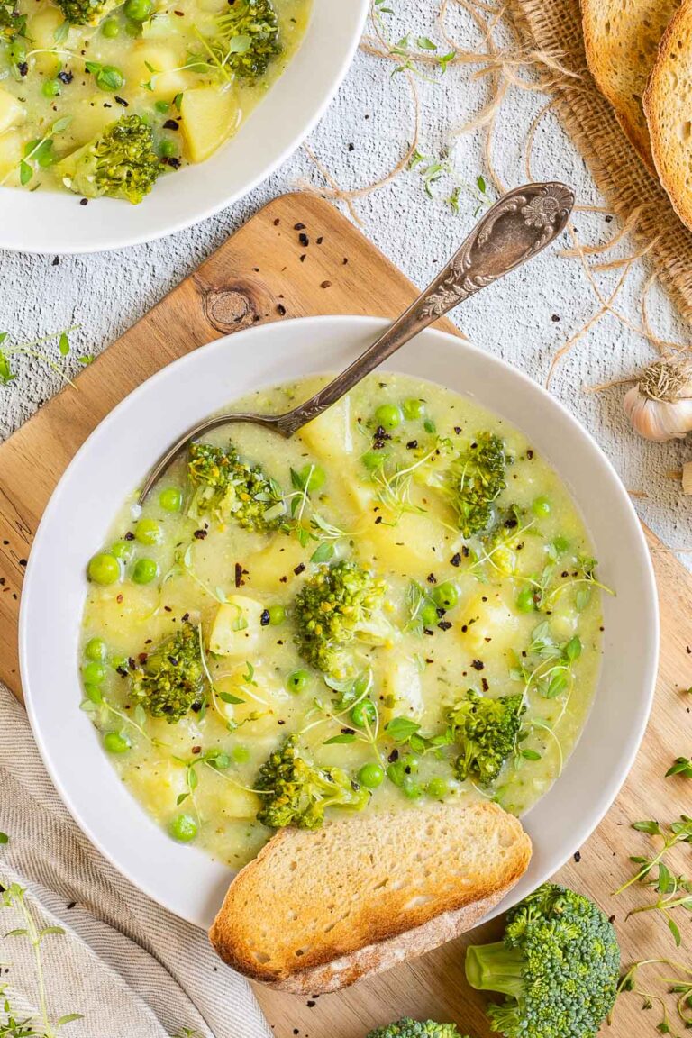 2 white plates of thick yellow stew with potatoes, broccoli, green peas and green herbs. A spoon is placed inside with a toasted slice of bread.