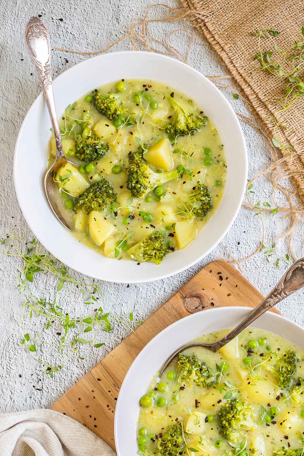 2 white plates of thick yellow stew with potatoes, broccoli, green peas and green herbs. A spoon is placed inside with a toasted slice of bread.