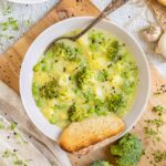 A white plate of thick yellow stew with potatoes, broccoli, green peas and green herbs. A spoon is placed inside with a toasted slice of bread.