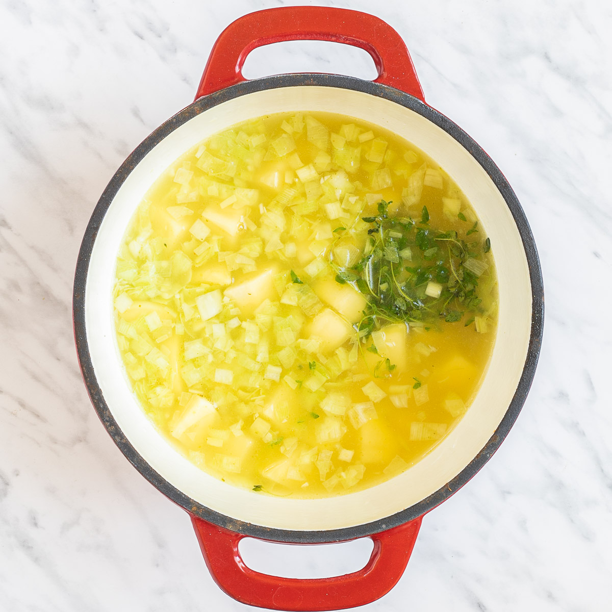 Red and white enameled Dutch oven with large potato chunks, leek slices and green herbs in a light yellow liquid.