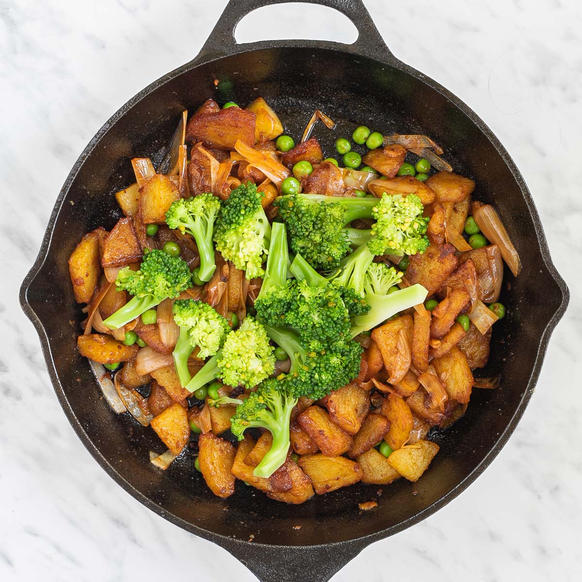 Skillet with potatoes, green peas, and caramelized onion slices topped with a heap of brocccoli florets.
