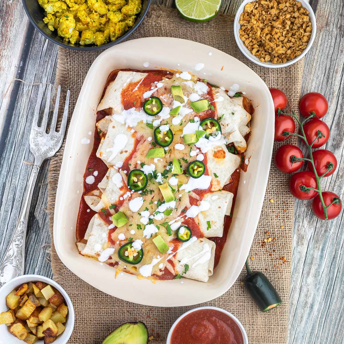 4 tortilla wraps topped with red sauce, melted cheese, sour cream, green chili peppers and fresh chopped herbs tucked closely together in a ceramic pan. Leftover filling ingredients are next to the pan in small bowls.