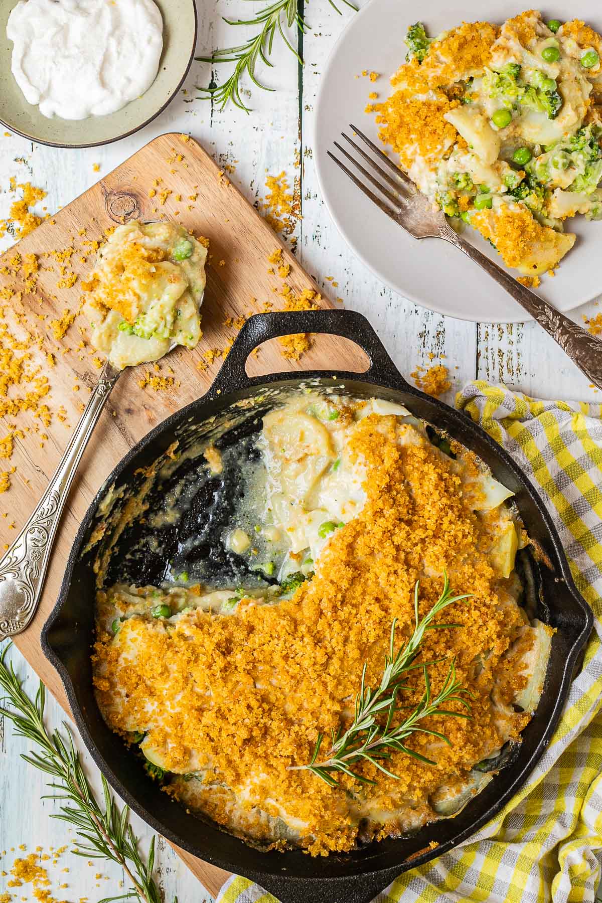 A black cast iron skillet with potato slices in a yellow thick sauce topped with yellow breadcrumbs. A part of it is missing and there is a gray plate with some food on it where you can see peas and broccoli as well. 