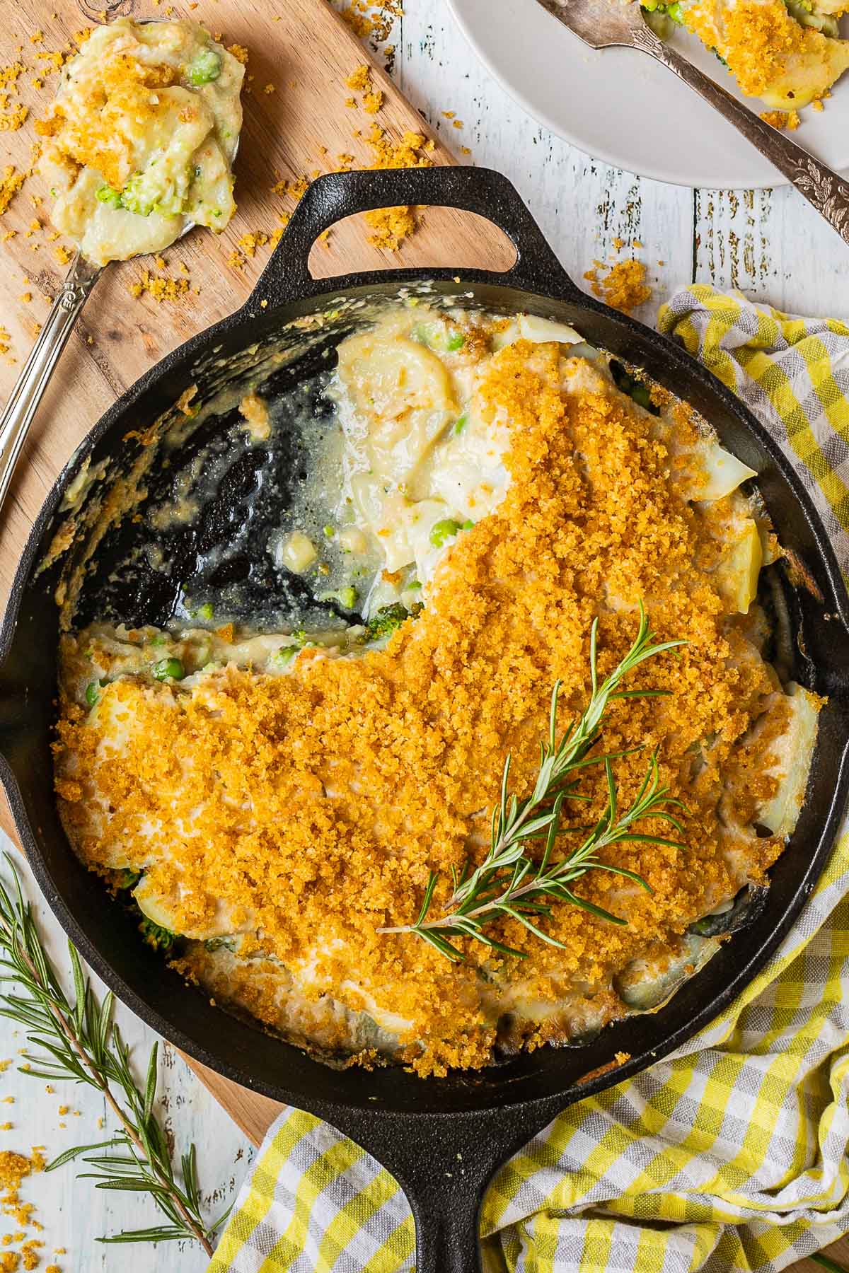 A black cast iron skillet with potato slices in a yellow thick sauce topped with yellow breadcrumbs. A part of it is missing and there is a large serving spoon next to it with some of the food from the skillet 