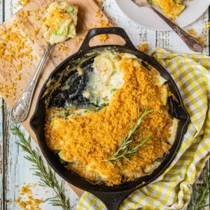 A black cast iron skillet with potato slices in a yellow thick sauce topped with yellow breadcrumbs. A part of it is missing and there is a large serving spoon next to it with some of the food from the skillet 