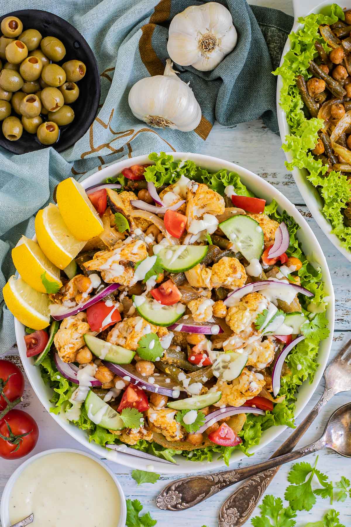 A large white bowl with green salad leaves topped with cauliflower florets, green beans, chickpeas, fresh tomatoes drizzled with a white sauce. Olives and more roasted veggies are next to it on small plates.