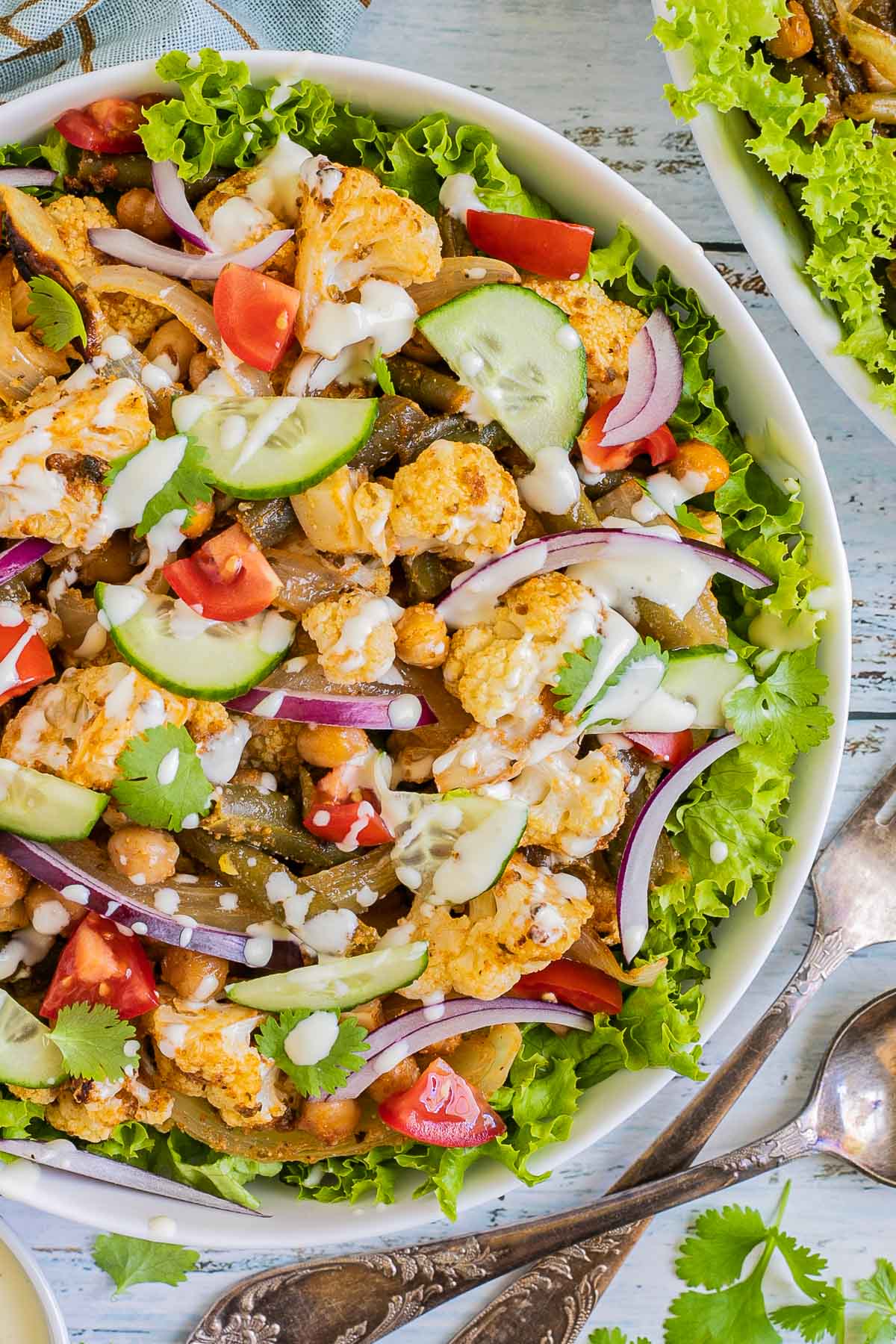 A large white bowl with green salad leaves topped with cauliflower florets, green beans, chickpeas, fresh tomatoes drizzled with a white sauce. Olives and more roasted veggies are next to it on small plates.