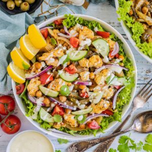 A large white bowl with green salad leaves topped with cauliflower florets, green beans, chickpeas, fresh tomatoes drizzled with a white sauce. Olives and more roasted veggies are next to it on small plates.