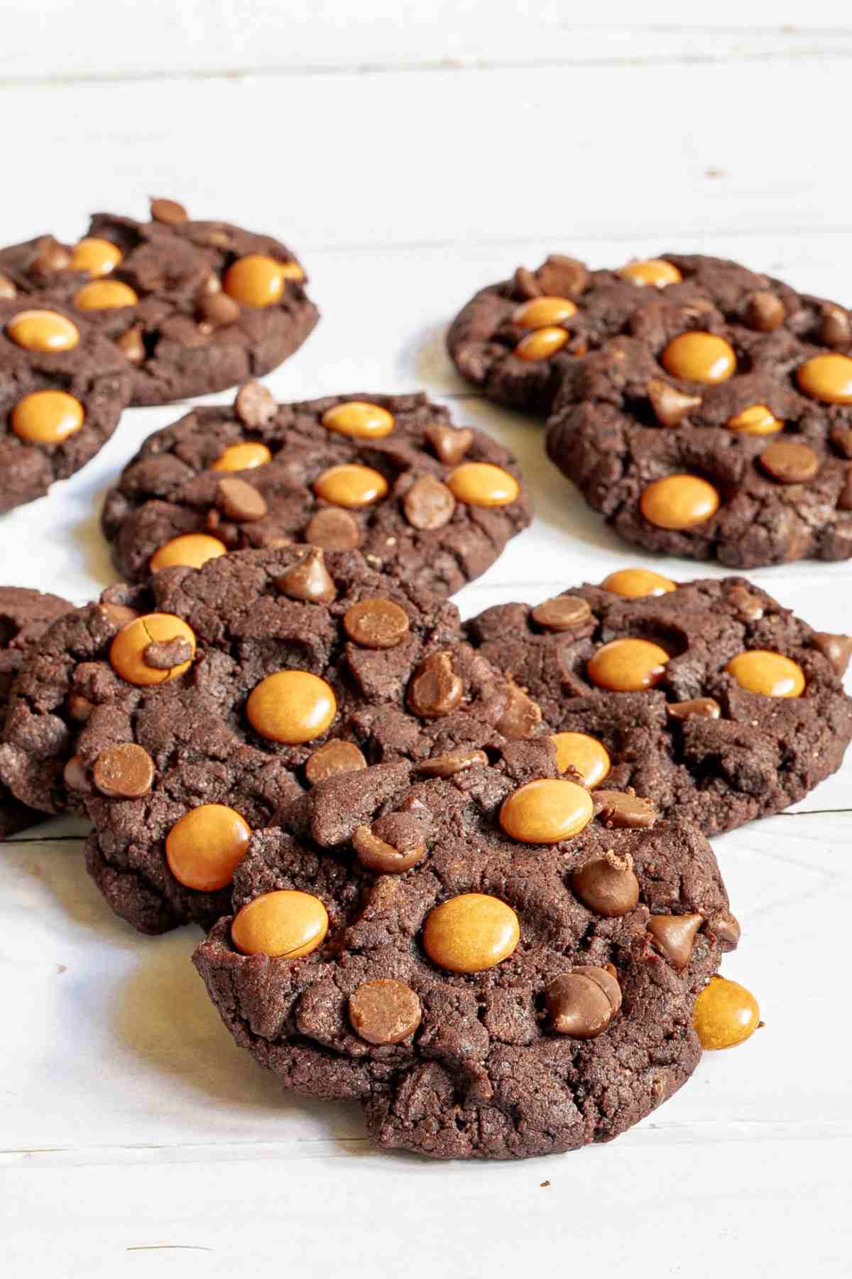 Dark brown cookies with orange m&ms, and chocolate chips on a white wooden surface. A hand is grabbing one.