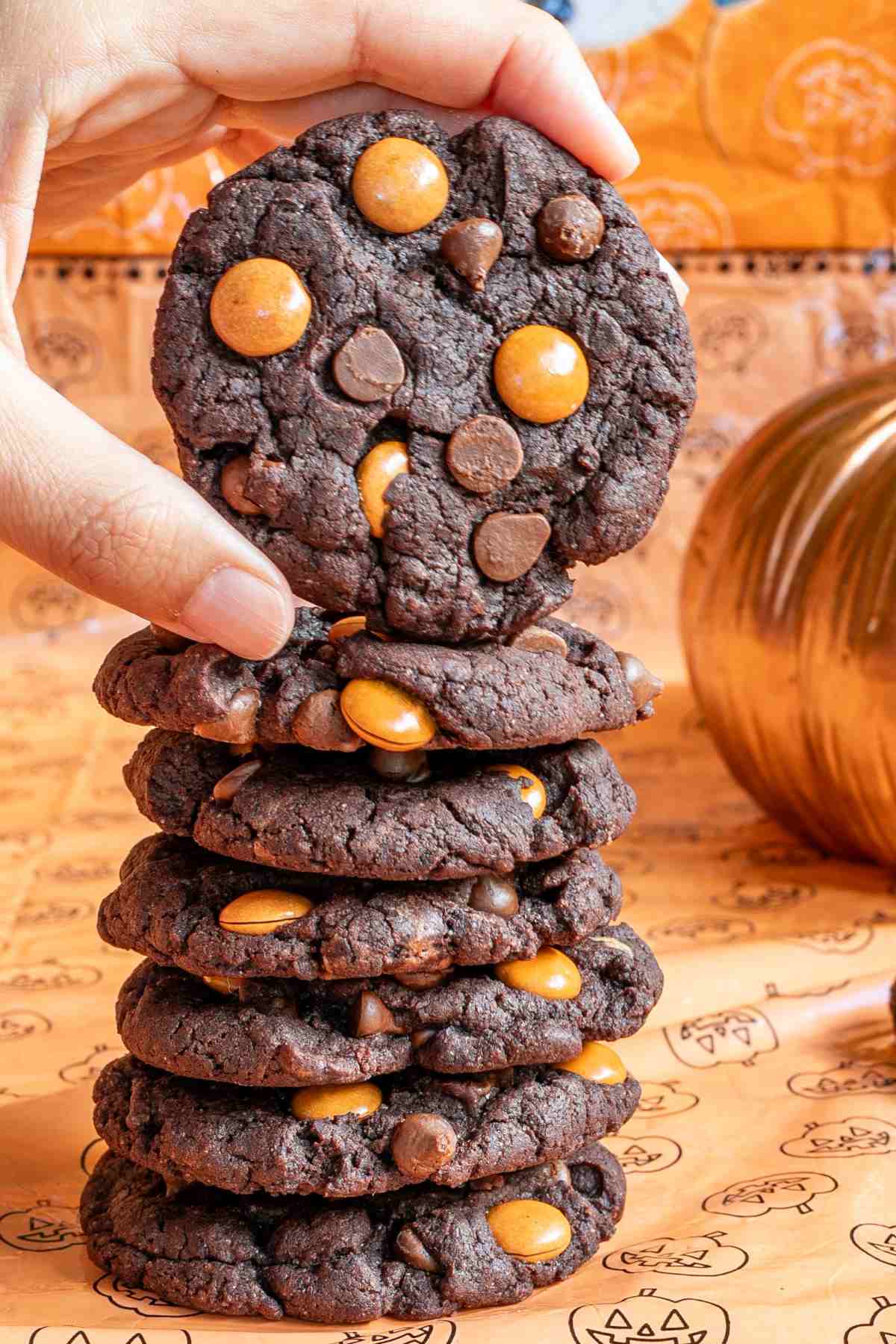 A stack of 6 dark brown cookies with orange m&ms, and chocolate chips on an orange surface with a pumpkin pattern. A hand is grabbing the top one.