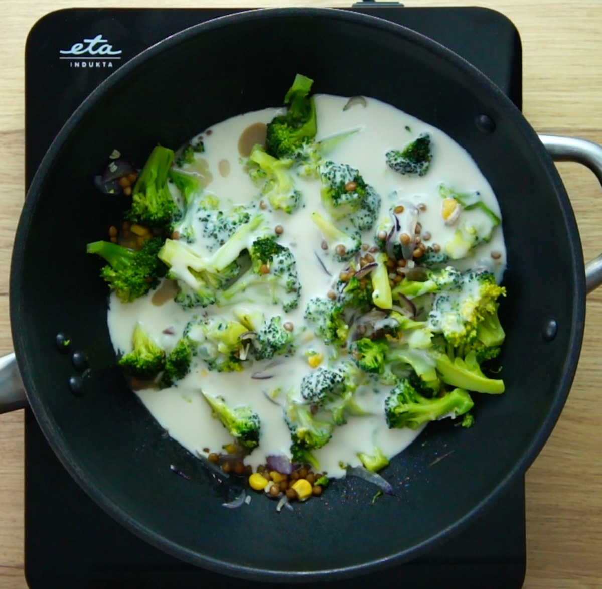 A wok with red onion slices, brown lentils, yellow corn, and broccoli florets. White sauce is poured on top of them.