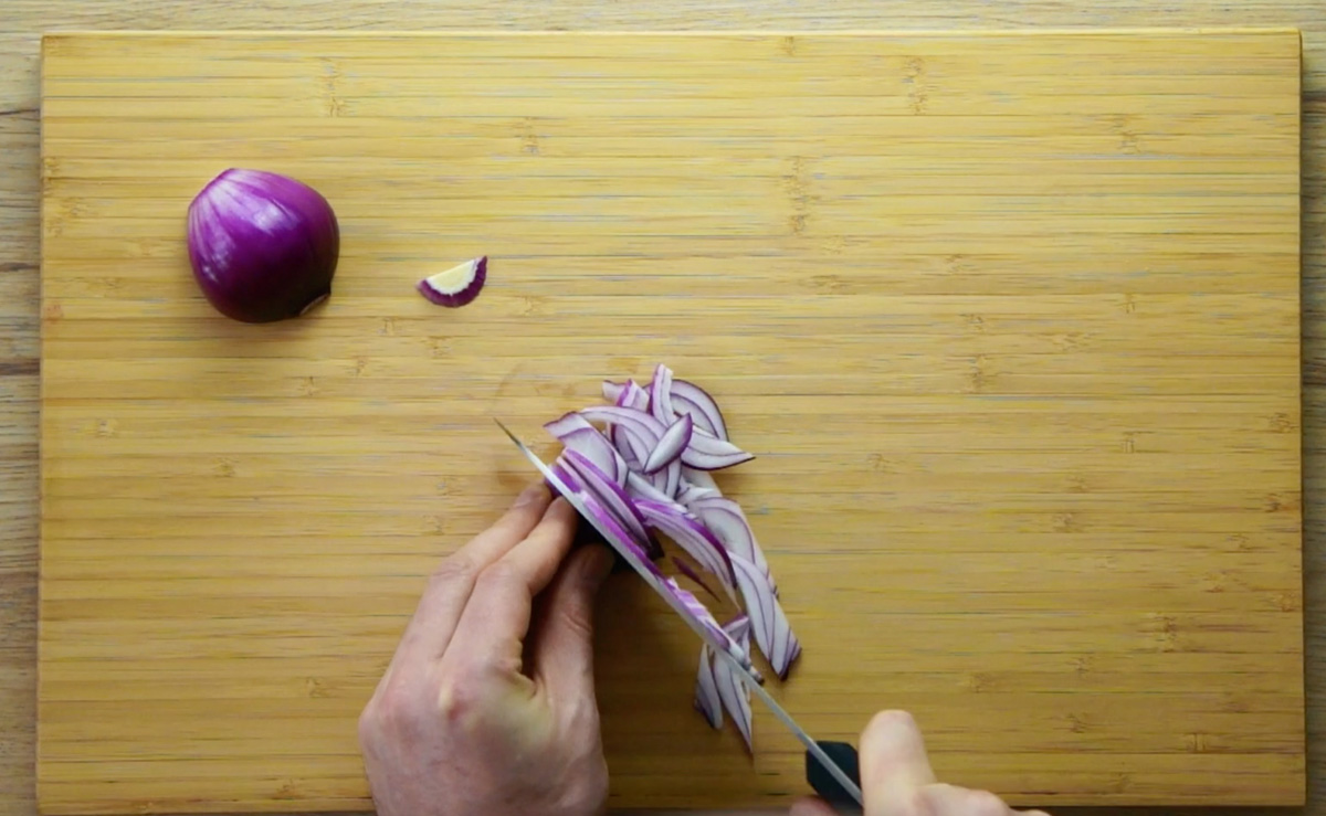 A wooden cutting board with half of a red onion. A hand is slicing the other half of the onion.