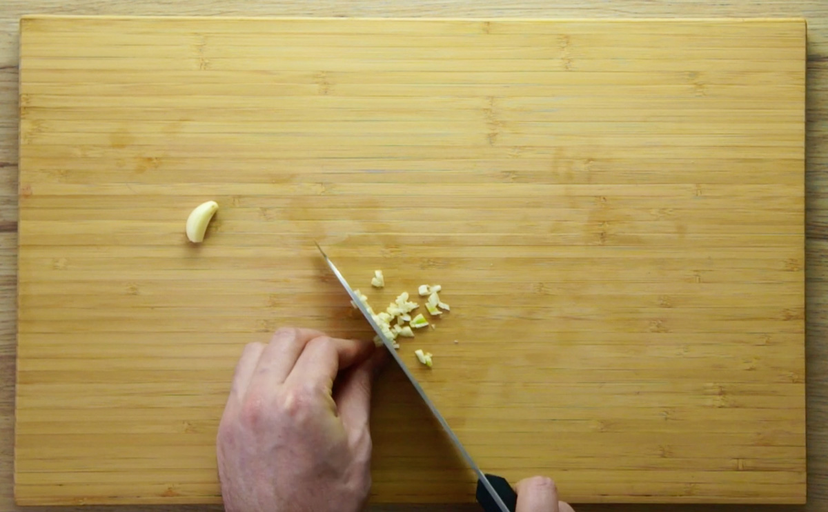 A wooden cutting board. A hand is cutting a garlic clove into very small pieces.