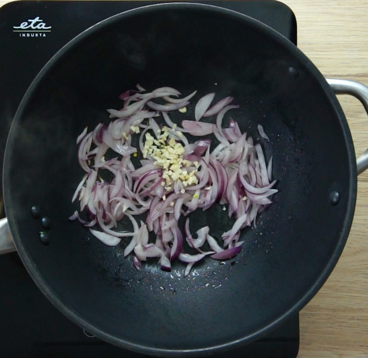 A wok with several red onion slices and a small heap of minced garlic.