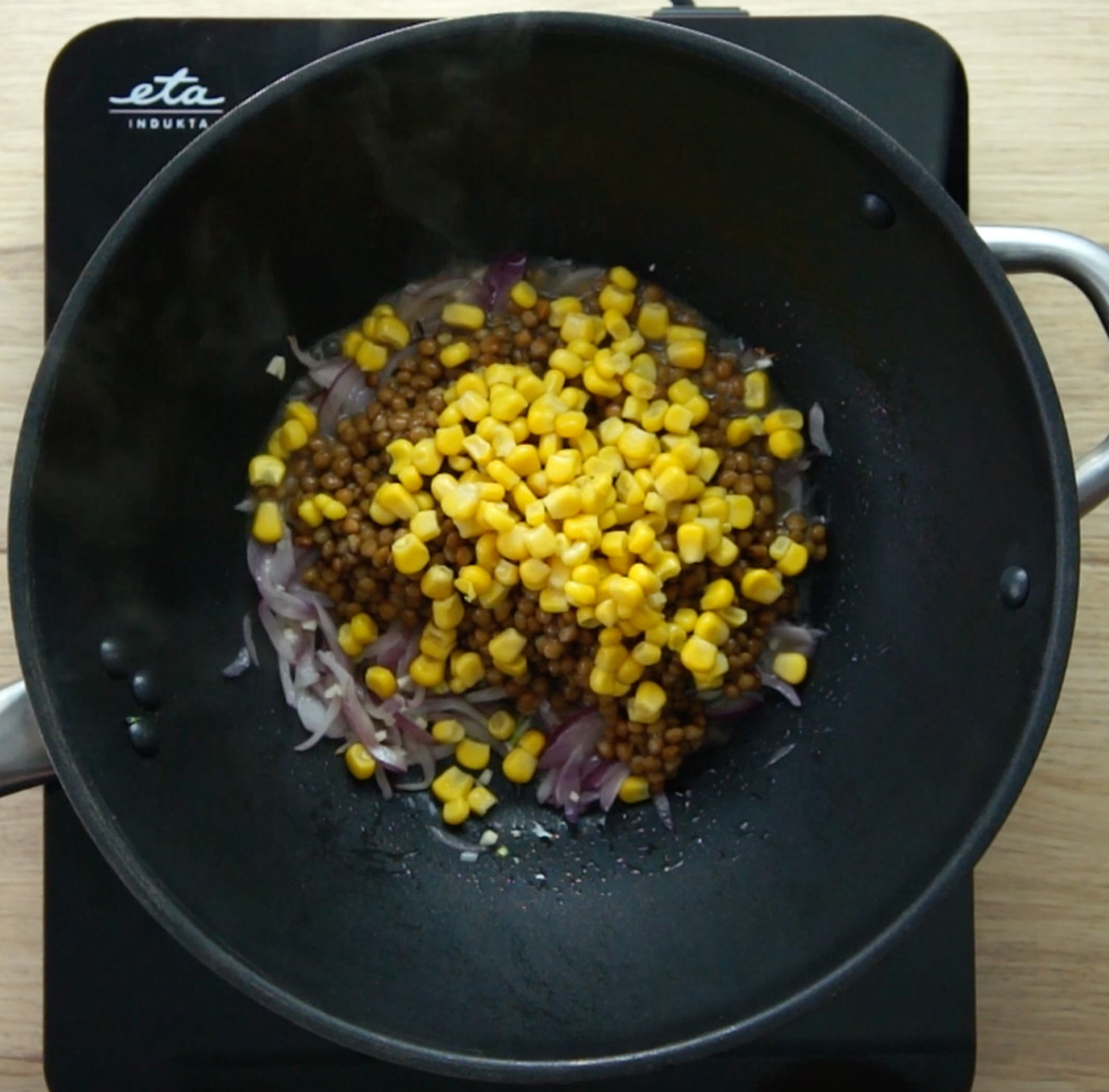 A wok with several red onion slices with a large heap of brown lentils and yellow corn.
