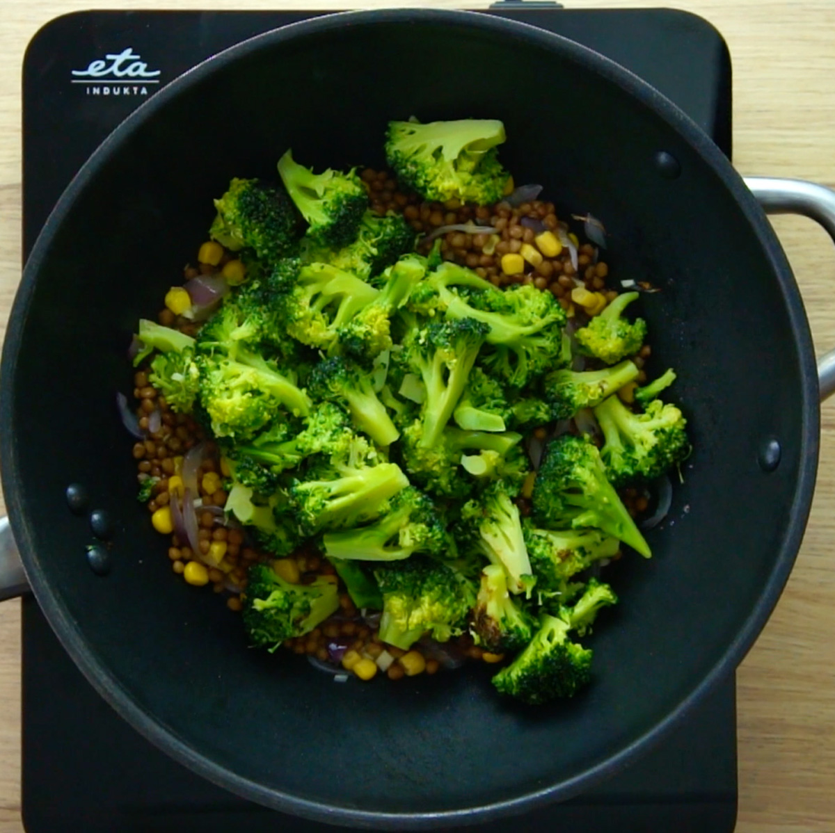 A wok with several red onion slices, brown lentils, and yellow corn with a lot of broccoli florets on top.