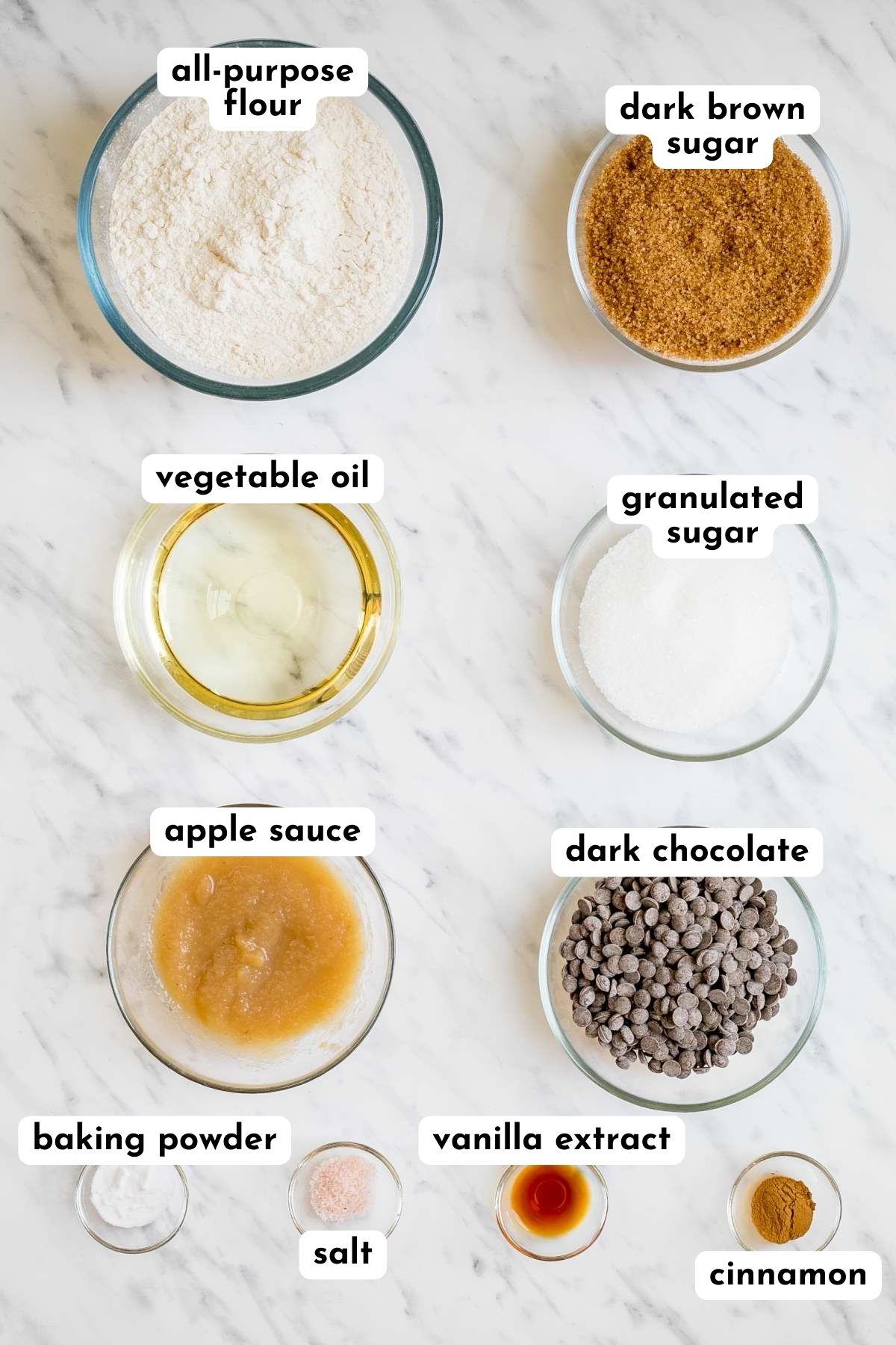 Ingredients of chocolate chip cookies in small glass bowls like flour, sugar, dark chocolate chips, apple sauce, oil, salt, cinnamon, vanilla extract, and baking soda.