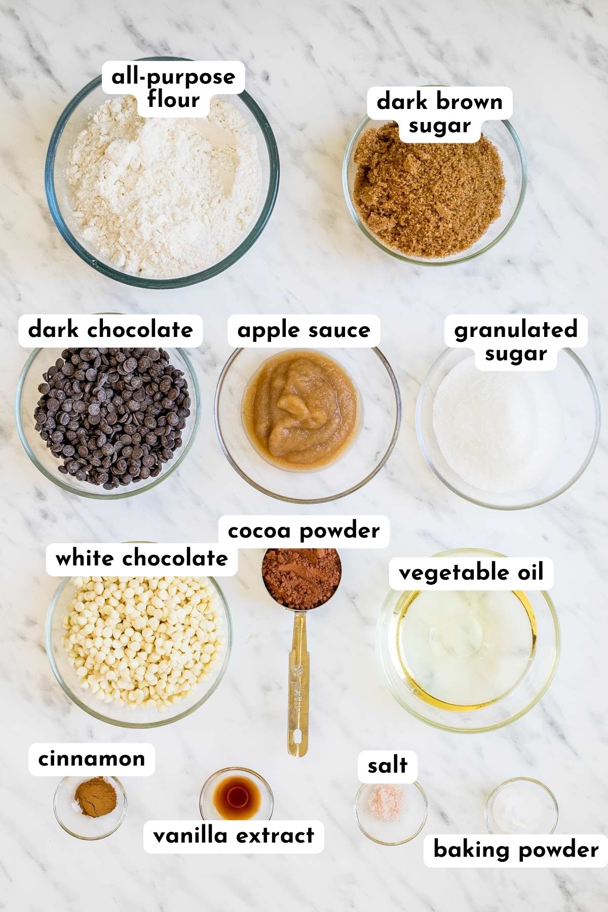 Ingredients of chocolate cookies in small glass bowls like flour, brown sugar, white sugar, cocoa powder, apple sauce, oil, salt, cinnamon, vanilla extract, baking soda, white and dark chocolate chips.